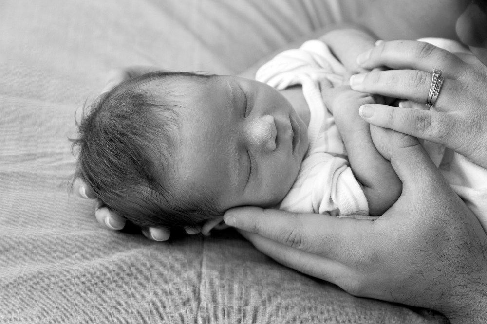 Black and white image of a newborn, sleeping, mum and dads hands surrounding him.