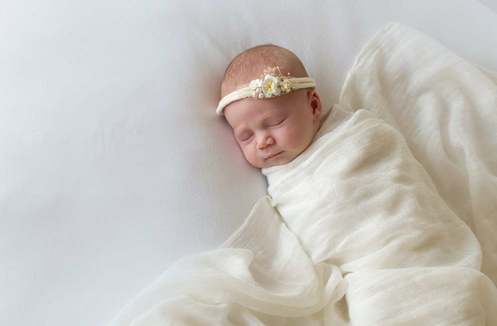 photograph of newborn baby during studio session. White on white with a small floral headband/tieback.