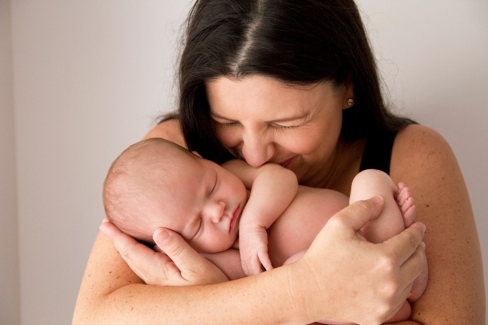 close up photograph of new mum cuddling and kissing her new baby. Baby asleep and curled up in mums arms.